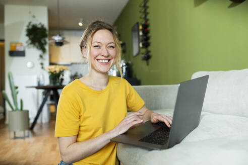 Glücklicher Freiberufler in gelbem T-Shirt bei der Arbeit mit Laptop zu Hause - MIKF00300