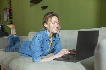 Freelancer in denim shirt working with laptop lying on couch - MIKF00303