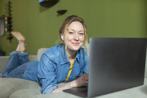 Freiberufler in Jeanshemd arbeitet mit Laptop auf der Couch liegend - MIKF00304