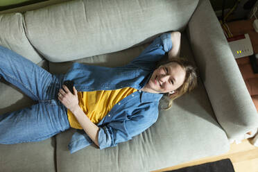 Young woman enjoying music with wireless in-ear headphones lying on couch - MIKF00307