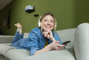 Nachdenkliche Frau, die mit Kopfhörern auf der Couch liegend Musik genießt - MIKF00313