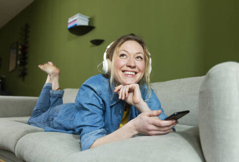 Thoughtful woman enjoying music with headphones lying on couch - MIKF00313