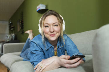 Smiling young woman listening music with headphones lying on couch - MIKF00314