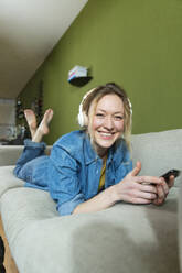 Lächelnde Frau hört Musik mit Kopfhörern auf der Couch liegend - MIKF00315