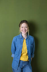 Smiling young woman in denim shirt standing in front of green wall - MIKF00319