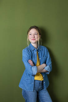 Nachdenkliche junge Frau mit verschränkten Armen lächelnd vor einer grünen Wand - MIKF00321