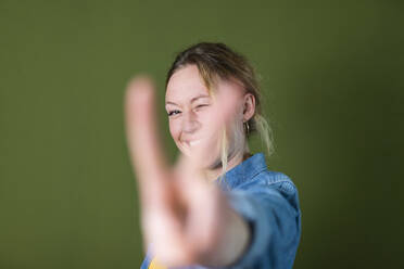 Smiling young woman gesturing peace sign in front of green wall - MIKF00325