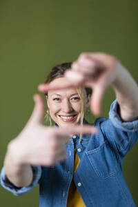 Lächelnde junge Frau macht Fingerrahmen vor grüner Wand - MIKF00329