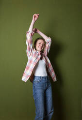 Smiling woman with arms raised standing in front of green wall - MIKF00331