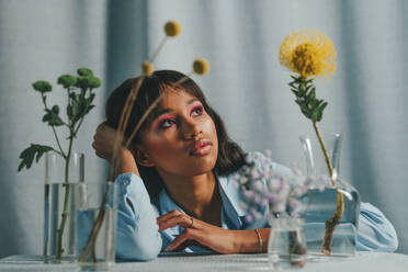 Thoughtful woman sitting at table with flowers in front of blue backdrop - YTF00800