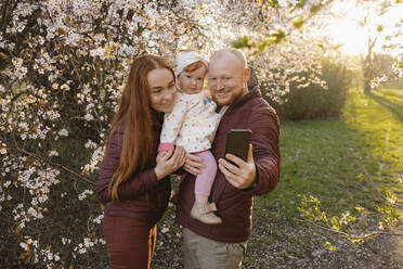 Eine Familie fängt einen Moment der Freude ein, als sie ein gemeinsames Selfie im Park macht, während ihre Tochter an einem Baum steht - VIVF00922