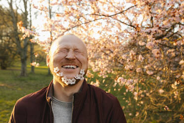 Glücklicher Mann mit Blumen im Bart in der Nähe eines Baumes im Park - VIVF00930