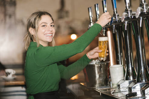 Happy young waitress filling beer glasses from tap at bar - EGAF02601