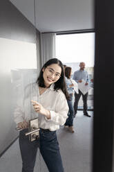 Porträt einer lächelnden jungen Geschäftsfrau mit Laptop und Kollegen im Hintergrund im Büro - KMKF01976