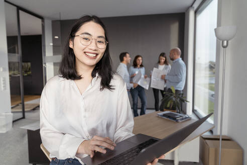 Porträt einer lächelnden jungen Geschäftsfrau mit Laptop und Kollegen im Hintergrund im Büro - KMKF01978