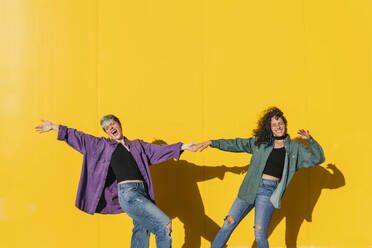 Cheerful lesbian couple holding hands in front of yellow wall - MGRF00930
