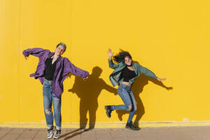 Glückliches lesbisches Paar springt vor einer gelben Wand an einem sonnigen Tag - MGRF00932