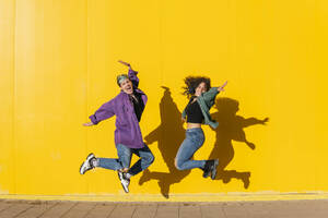 Fröhliches lesbisches Paar springt vor einer gelben Wand - MGRF00933