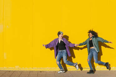 Cheerful lesbian couple running together in front of yellow wall - MGRF00937