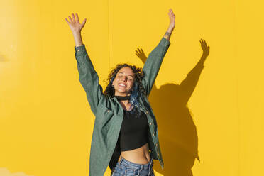 Carefree woman with arms raised in front of yellow wall - MGRF00940
