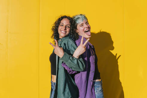 Cheerful lesbian women showing peace sign in front of yellow wall - MGRF00943