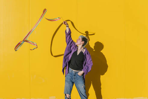 Lesbian woman waving rainbow colored ribbon in front of yellow wall - MGRF00948