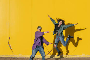 Happy woman with lesbian friend jumping in front of yellow wall - MGRF00950