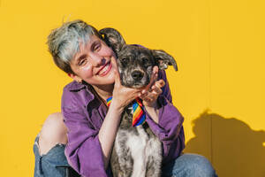 Happy lesbian woman with dog in front of yellow wall - MGRF00958