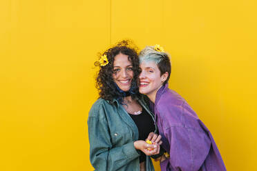 Happy lesbian couple with flowers on hair standing in front of yellow wall - MGRF00970