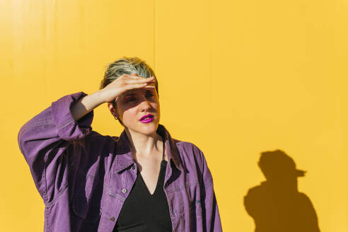 Frau schützt ihre Augen vor Sonnenlicht vor einer gelben Wand - MGRF00973