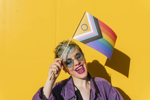 Glückliche lesbische Frau mit Regenbogenfahne vor einer gelben Wand - MGRF00977