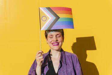 Happy lesbian woman with eyes closed holding rainbow flag in front of yellow wall - MGRF00981