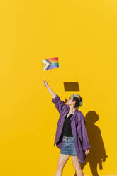 Lesbische Frau mit mehrfarbiger Flagge vor gelber Wand stehend - MGRF00985