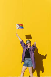 Lesbische Frau mit Friedenszeichen und Regenbogenflagge vor einer gelben Wand - MGRF00986