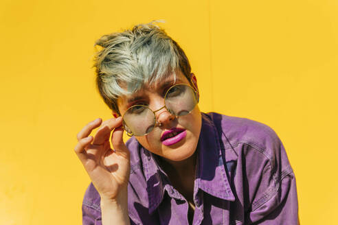 Woman with dyed hair holding sunglasses in front of yellow wall - MGRF00998