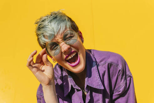 Carefree woman holding sunglasses and screaming in front of yellow wall - MGRF00999