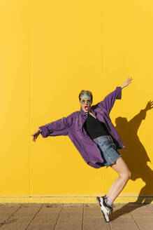 Carefree woman with arms outstretched dancing in front of yellow wall - MGRF01001
