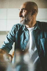 Lächelnder reifer Mann mit rasiertem Kopf und grauem Bart sitzt in der Küche - JOSEF19025