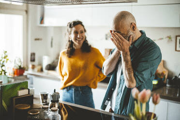 Mann, der sein Gesicht mit den Händen bedeckt, steht in der Küche neben einer lachenden Frau - JOSEF19027