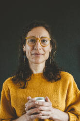 Smiling mature woman wearing eyeglasses holding coffee cup against black background - JOSEF19040