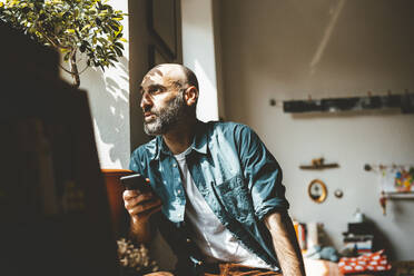 Contemplative man holding smart phone at home - JOSEF19052