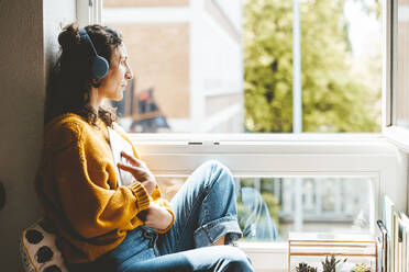 Thoughtful woman wearing headphones sitting near window at home - JOSEF19063