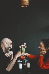 Happy couple giving fist bump sitting in front of black wall - JOSEF19080