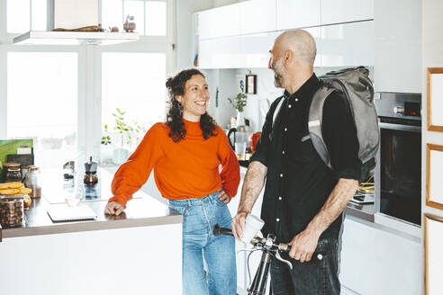 Man with bicycle talking to woman standing in kitchen at home - JOSEF19102