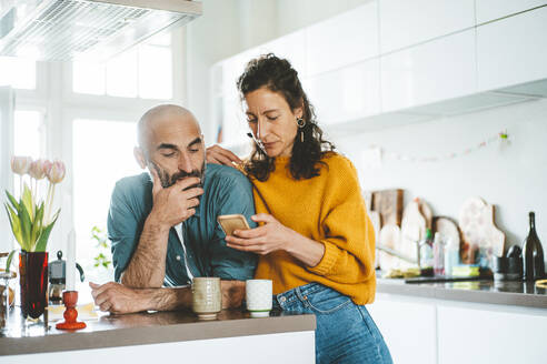 Woman sharing smart phone with man leaning on kitchen counter at home - JOSEF19108