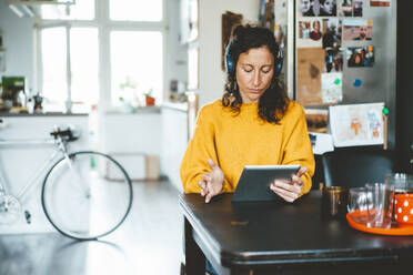 Woman wearing wireless headphones using tablet PC sitting at home - JOSEF19118