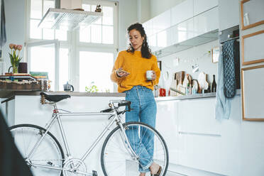 Frau, die ein Smartphone benutzt, stehend neben einem Fahrrad in der Küche zu Hause - JOSEF19121