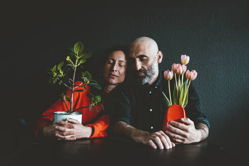 Mature couple with eyes closed holding potted plant and vase in front of black wall - JOSEF19141