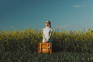 Frau mit altem Koffer steht inmitten von Pflanzen in einem Rapsfeld - VSNF00803