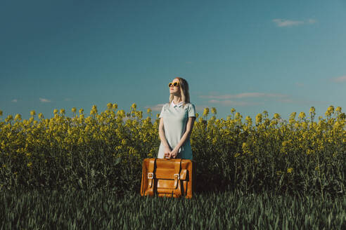 Woman with vintage suitcase standing amidst plants in rapeseed field - VSNF00803